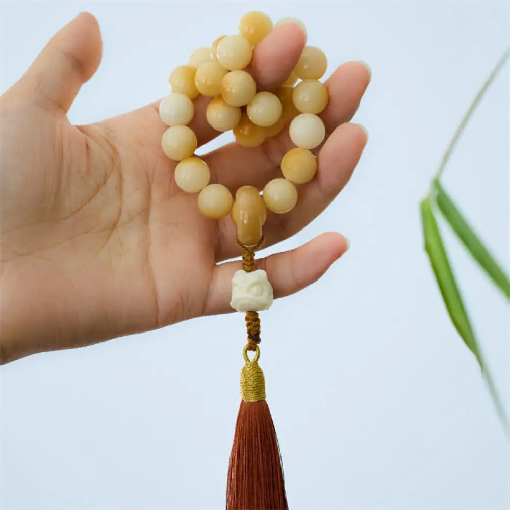 A close-up of the BWOOOH Natural Bodhi Seed Ivory Fruit Dancing Lion Charm Harmony Tassel Wrist Mala, featuring 12mm dark-speckled Bodhi Seed beads, smooth Ivory Fruit accents, and a detailed golden Dancing Lion charm with a vibrant harmony tassel, set against a muted, calming background.